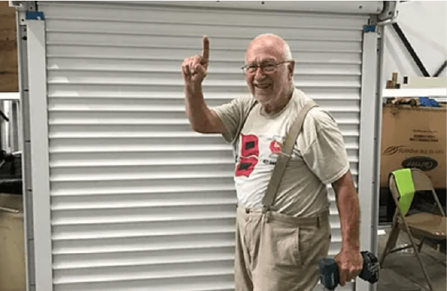 An older man is standing in front of a garage door holding a drill.