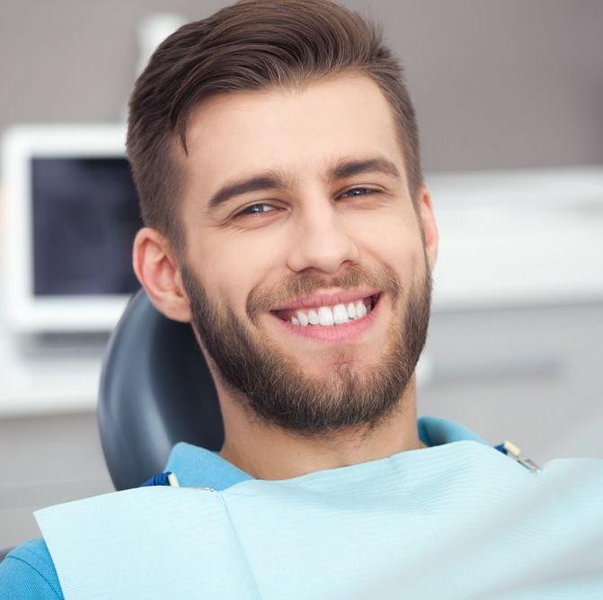 Man smiling in a dentist's chair, wearing a bib, blue shirt, clean white teeth.