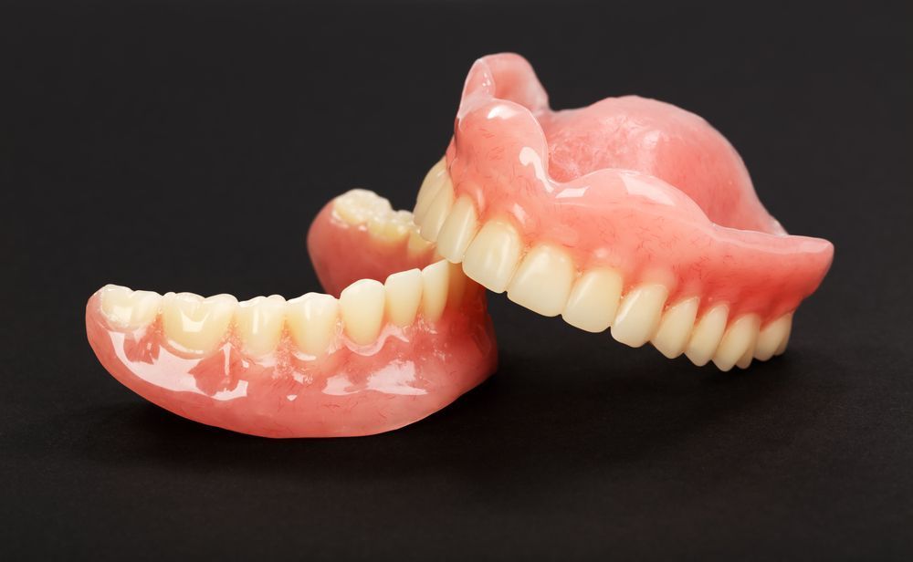 Dentures, top and bottom, on a black surface. The dentures have white teeth and pink gums.