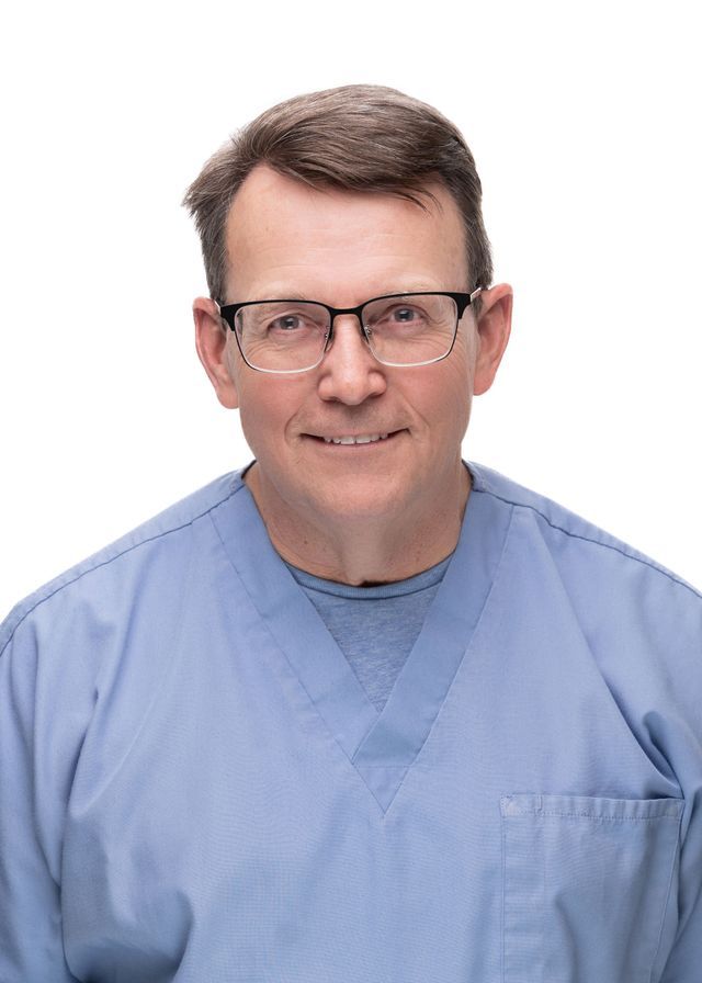 Man in blue scrubs and glasses smiling at the camera, against a white background.