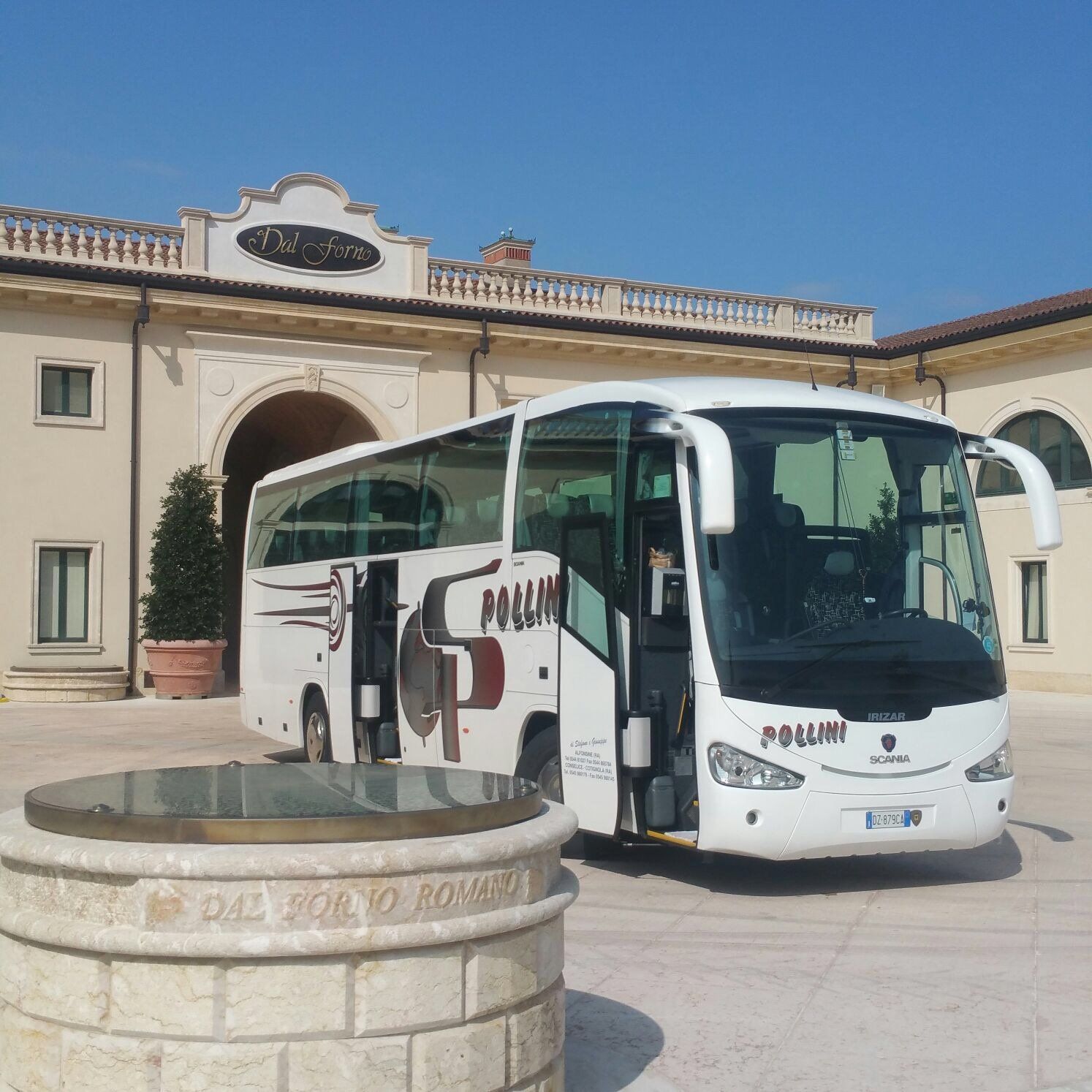 Un autobus turistico bianco parcheggiato fuori da un edificio con ingressi ad arco. In primo piano c'è un pozzo.