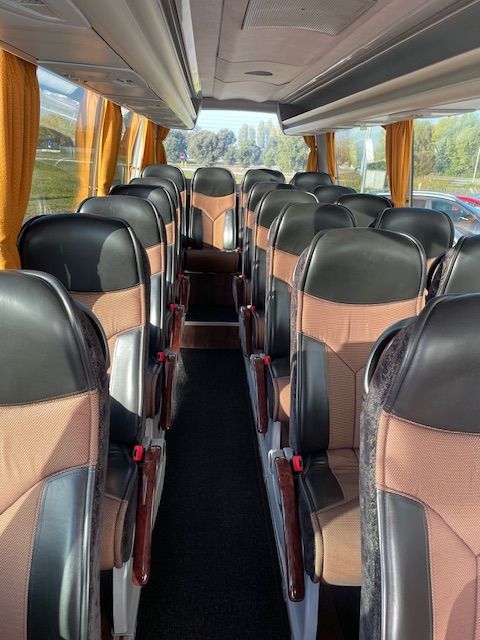 Interno di un autobus turistico con file di sedili neri e beige, corridoio centrale e finestrini con tende gialle.