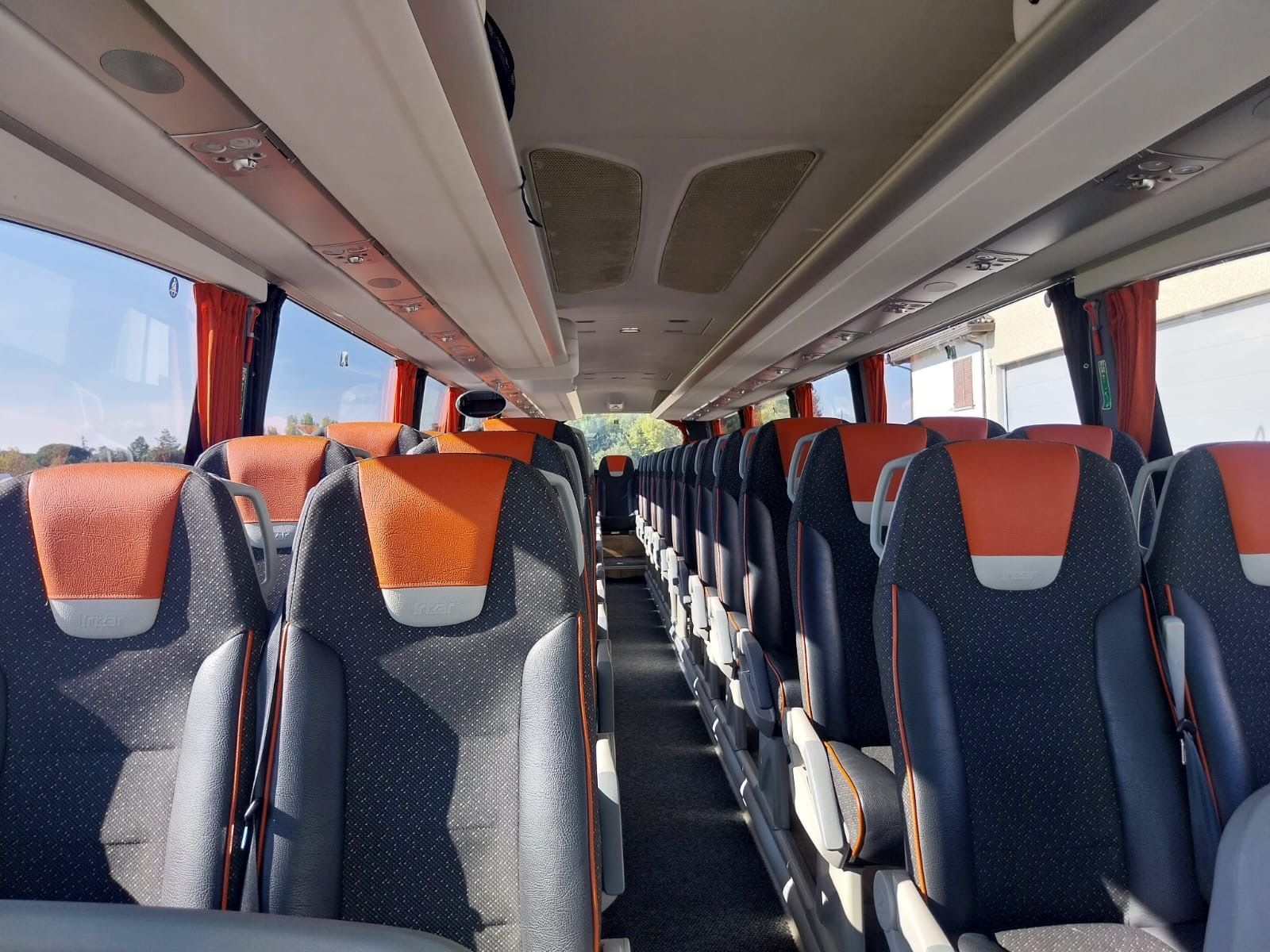 Interno di un autobus, file di sedili con poggiatesta arancioni e rivestimenti grigi, finestrini con tendine rosse.