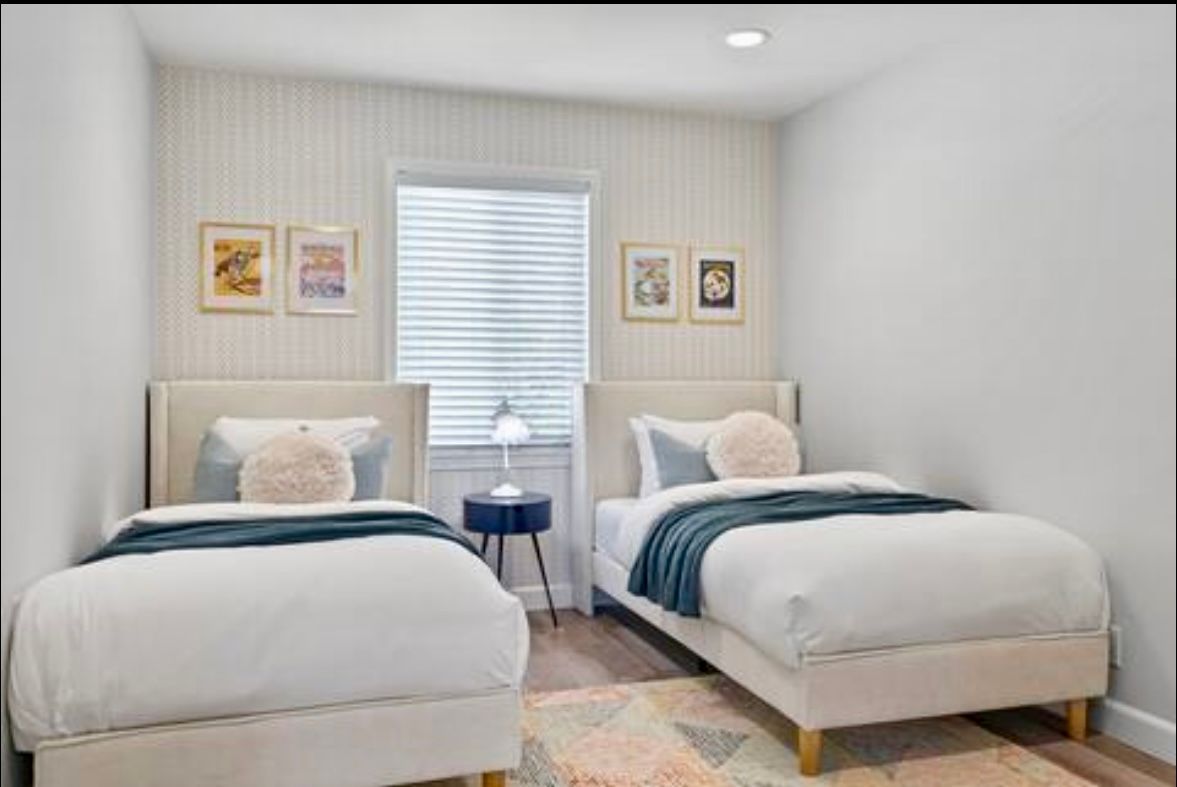 Two twin beds in a bedroom with artwork, a window, a side table, and a rug.