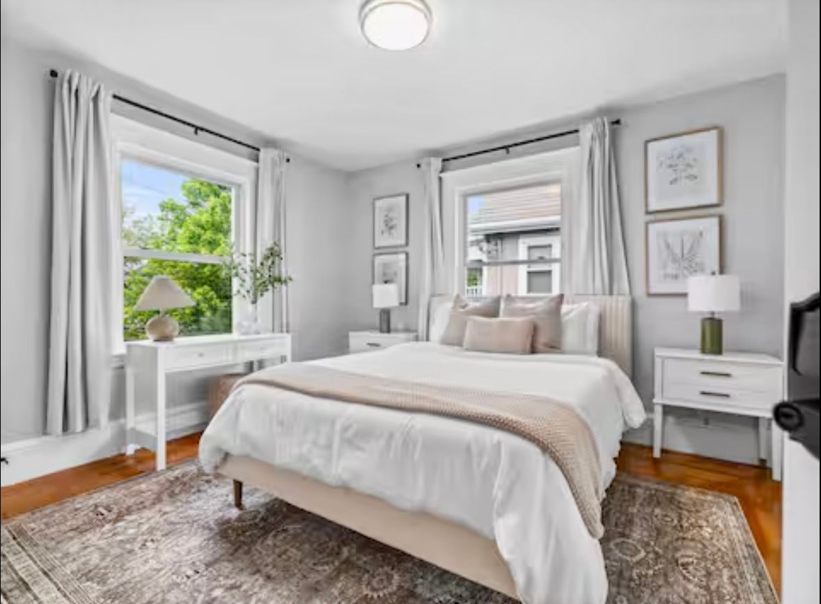 Bedroom with bed, desk, two windows, art, rug, white, gray, beige.
