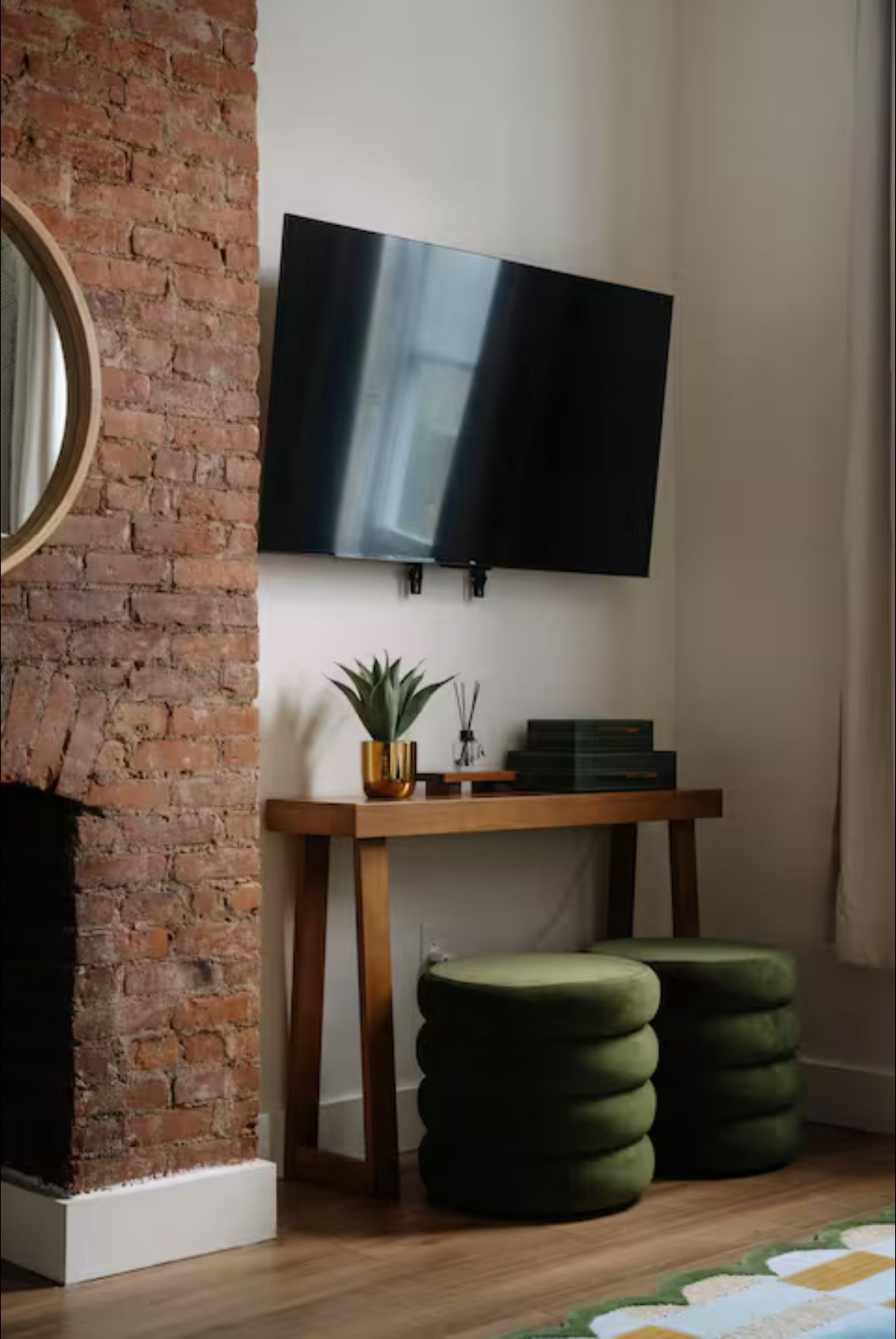 Living room corner with mounted TV, wooden console table, and two green ottomans. Brick wall and round mirror on the left.