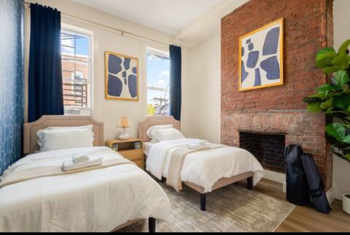 Bedroom with two beds, two windows, art, and exposed brick fireplace.