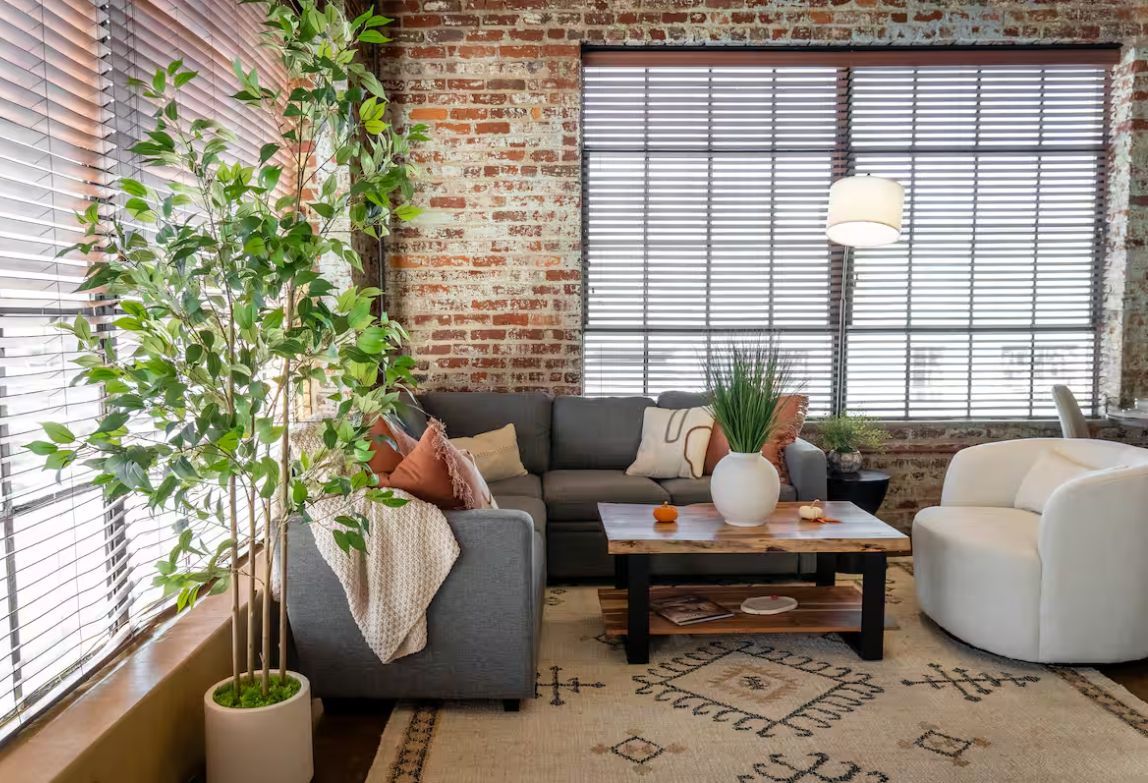 Living room with gray sectional, white chair, brick wall, and large window blinds.