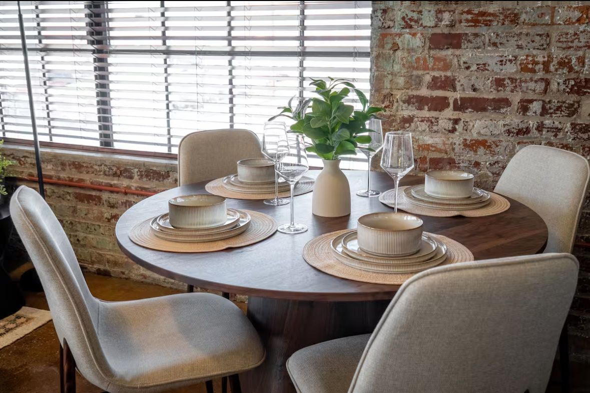 Round wooden dining table set for four, with cream chairs, against a brick wall and window.