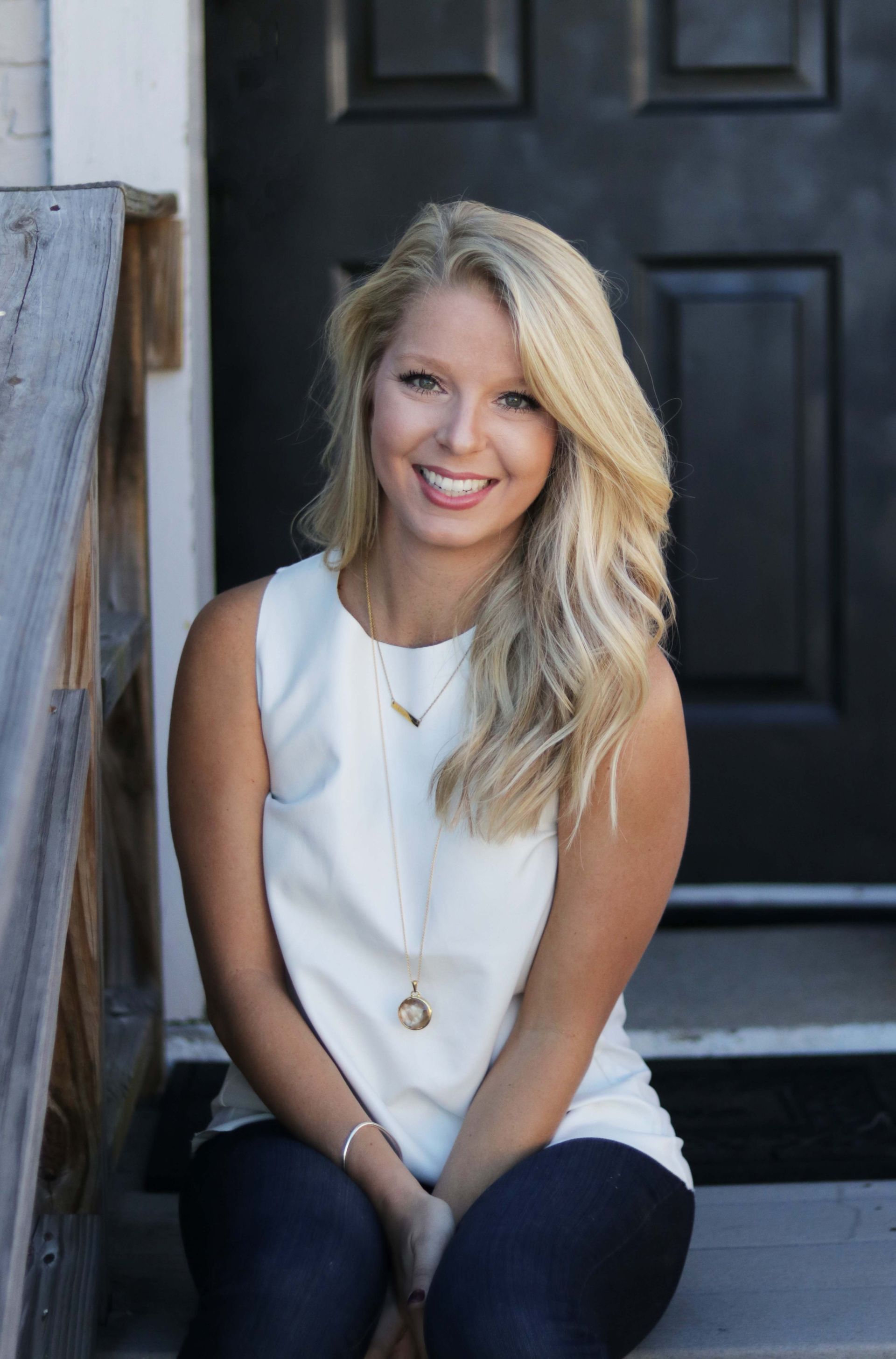 Blond woman smiling, sitting on steps, wearing a white top and dark jeans, in front of a black door.