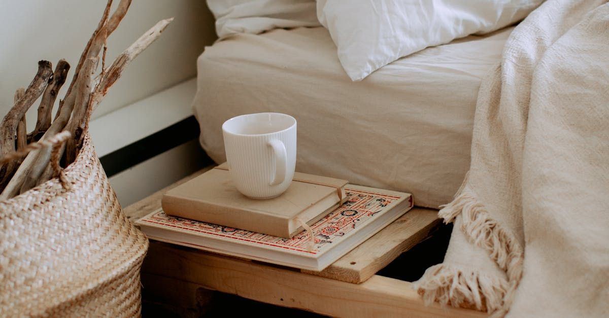 Woven basket with driftwood, books, and a mug on a bedside table beside a bed with white linens.