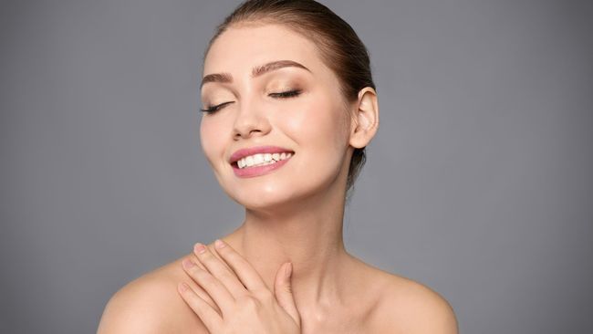 A woman is smiling with her eyes closed and touching her neck.