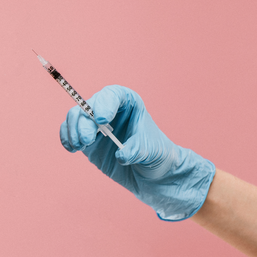 A person wearing blue gloves is holding a syringe on a pink background.