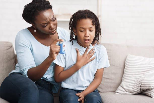 Woman helping child with an asthma inhaler on a couch; indoors, showing distress.