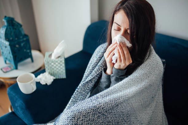 Woman on a blue sofa, wrapped in a blanket, blowing her nose with a tissue; sick.
