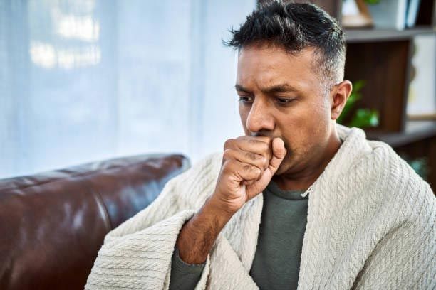 Man coughing, wrapped in blanket; appears to be sick, indoors.