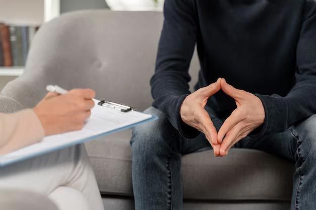 A person in a counseling session, with hands clasped, sitting on a couch while therapist takes notes.