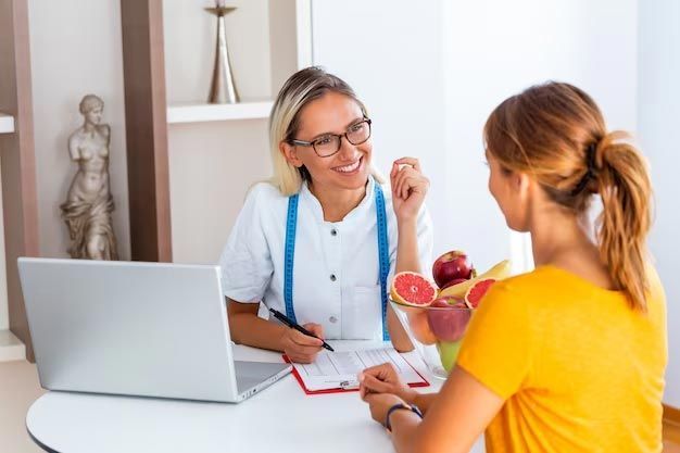 Nutritionist consulting with a patient, smiling, taking notes, with fruits on the table.