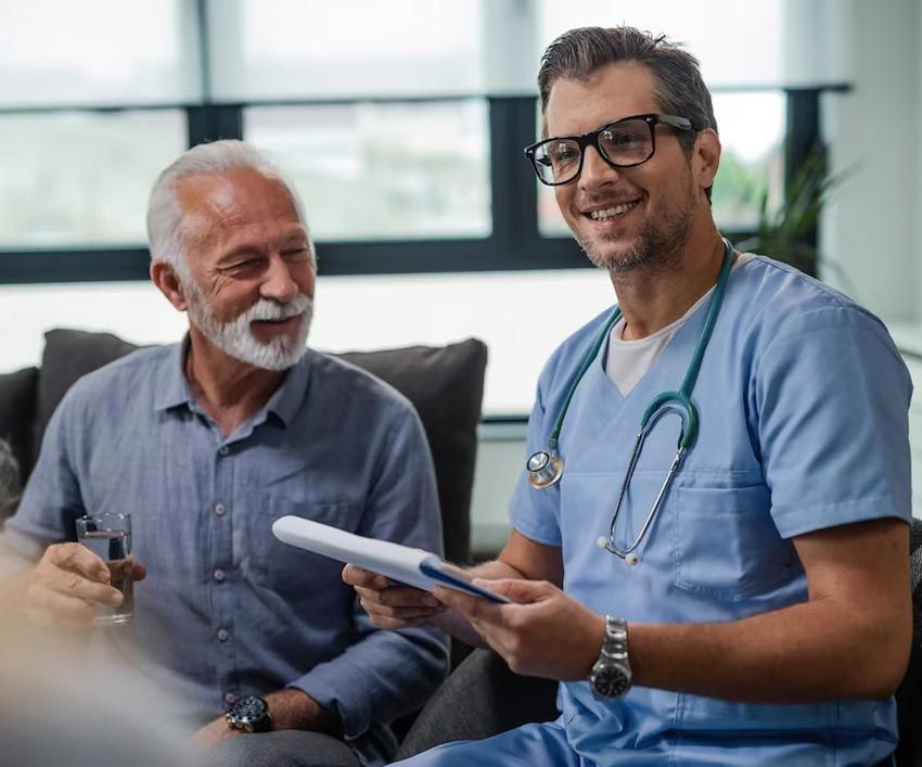 A smiling doctor in scrubs reviews a tablet with an elderly man in a home setting.