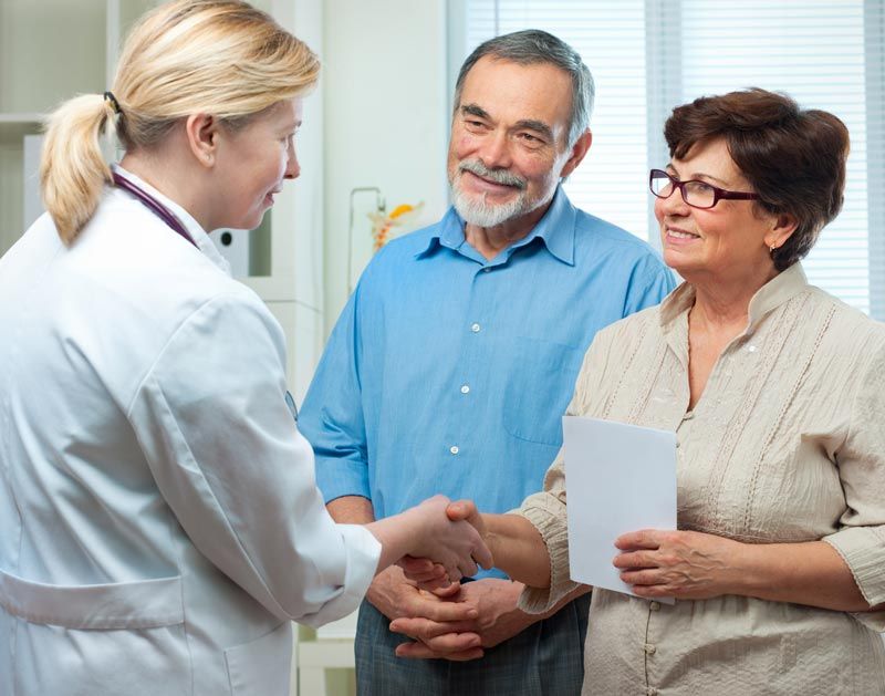 Doctor shaking hands with an older woman, man watches in a bright office.