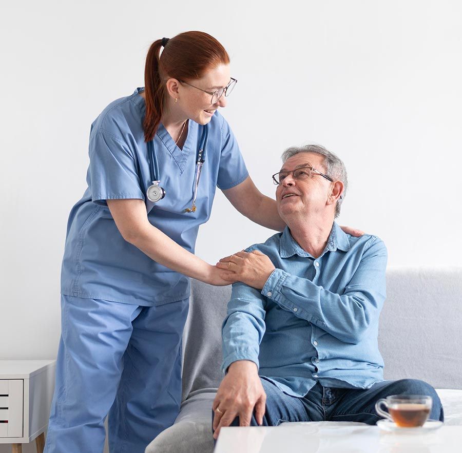 Nurse in blue scrubs comforts elderly man, indoors, with a cup of tea.