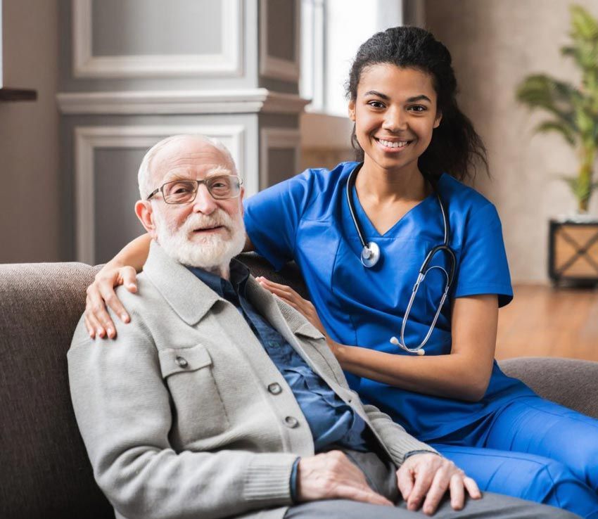 A smiling Black healthcare worker with stethoscope puts her arm around an elderly white man on a couch.