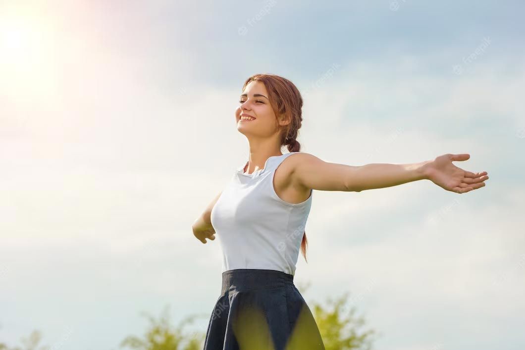 Woman with arms outstretched, smiling in a sunny outdoor setting.