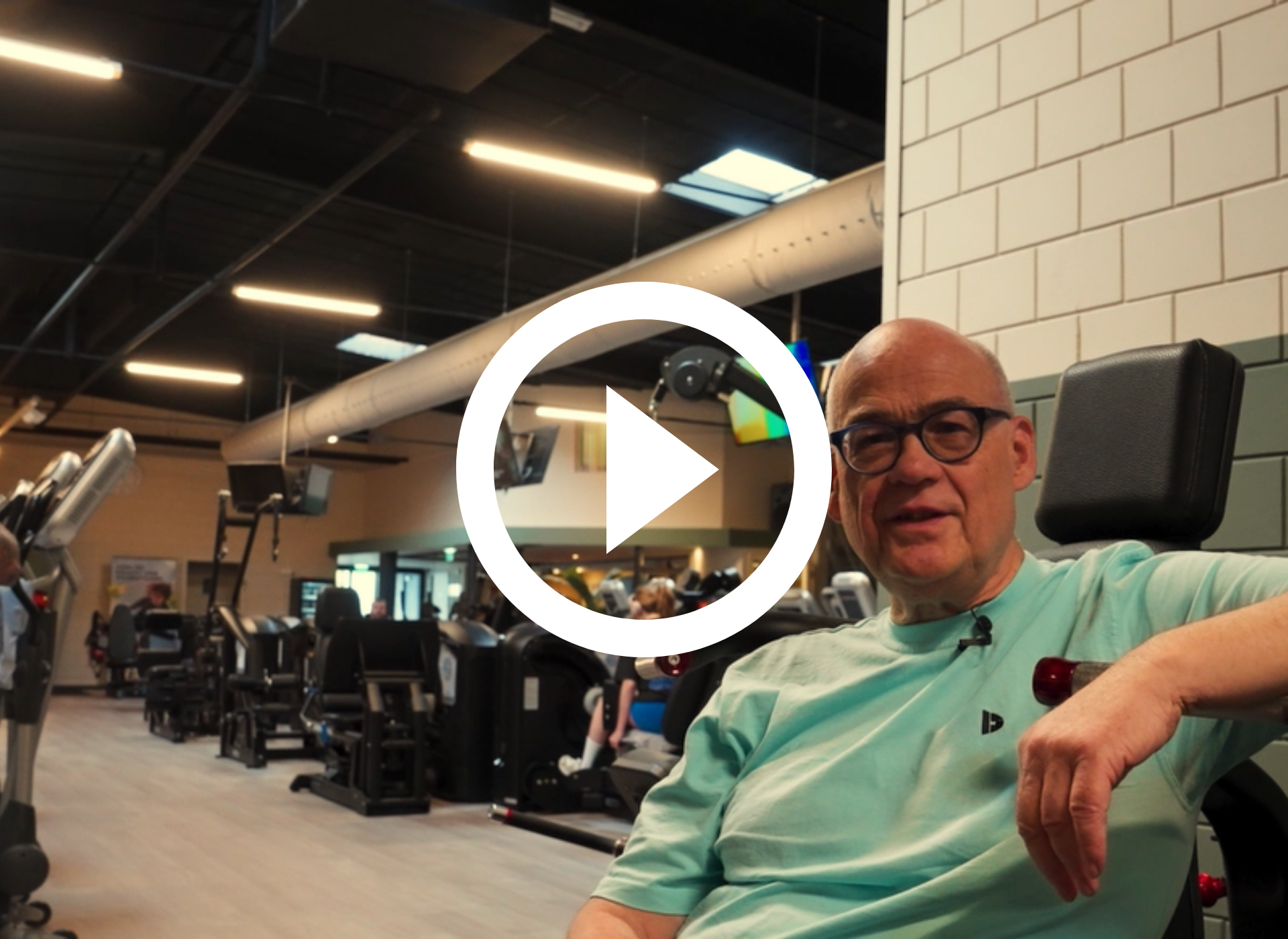 Een man zit in een sportschool, draagt ​​een bril en een mintgroen shirt, met fitnessapparaten op de achtergrond.