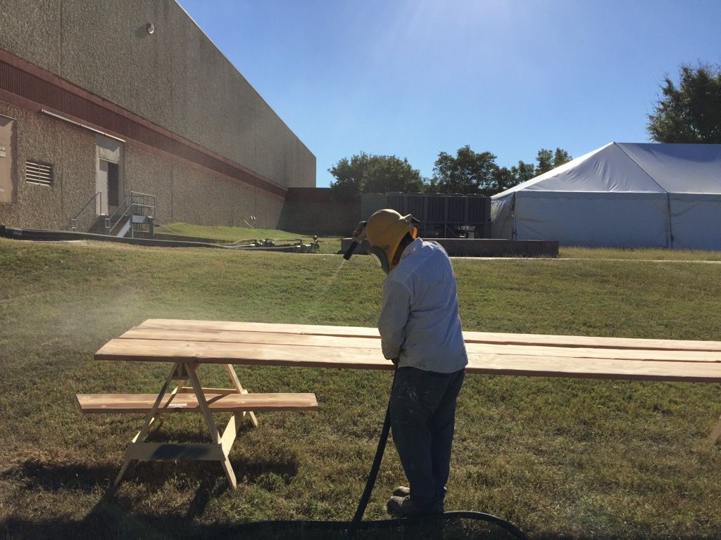 A man is spray painting a wooden picnic table in a field.
