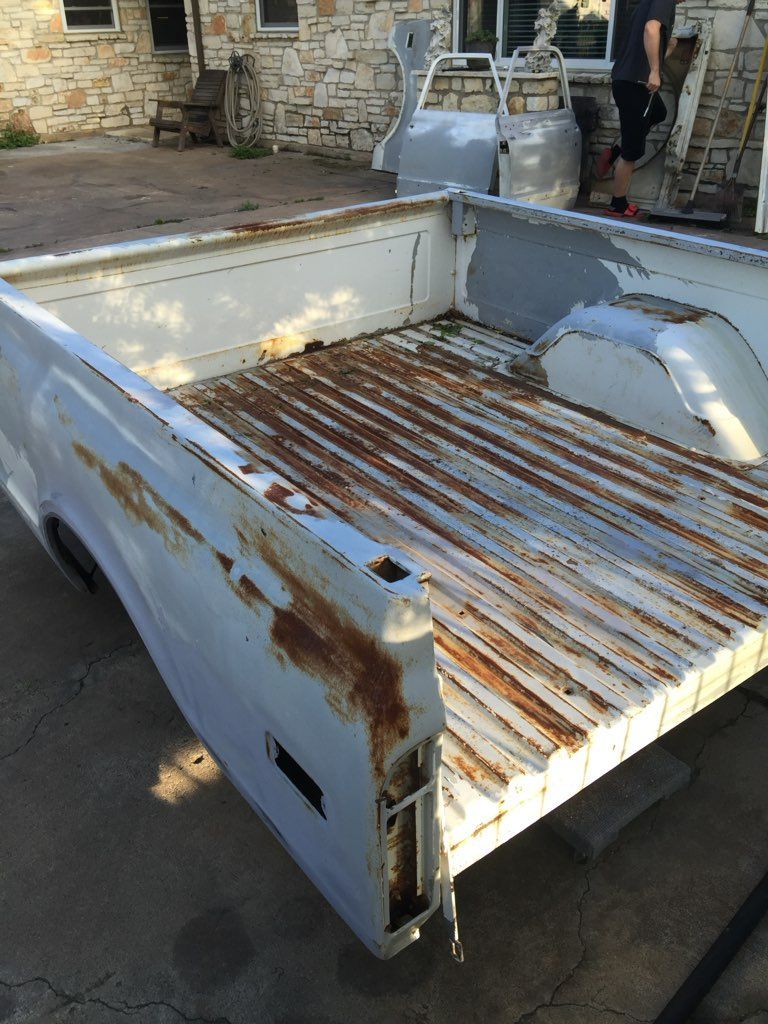 A white truck with a rusty bed is parked in front of a stone building.
