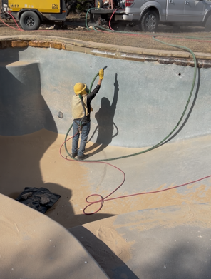 Person in protective gear sandblasting a drained swimming pool. Equipment and truck visible.