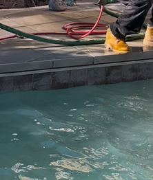 Person spraying water near a pool's edge, wearing work boots. Hoses and stone surround the pool.