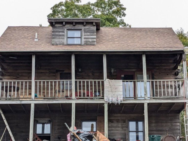 Two-story wooden cabin with covered balconies, brown logs, and a dormer window.