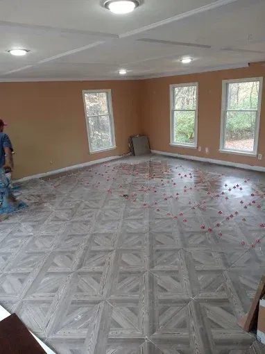 Room with tiled floor in progress; man working on floor; peach walls, white trim, three windows.