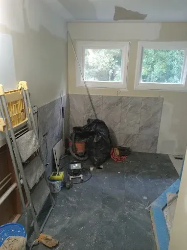 Bathroom under renovation; gray tile, ladder, and construction debris. Two windows with green trees in the background.