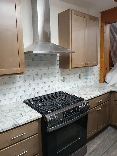 Kitchen with light brown cabinets, gas stove, stainless steel range hood, and white countertop with blue patterned backsplash.