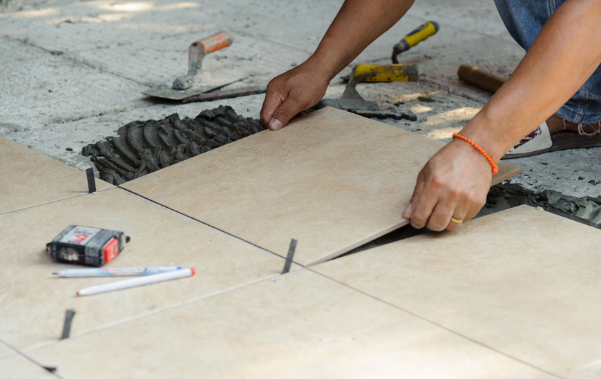 Person installing beige tiles outdoors, using tools, spacers, and mortar.