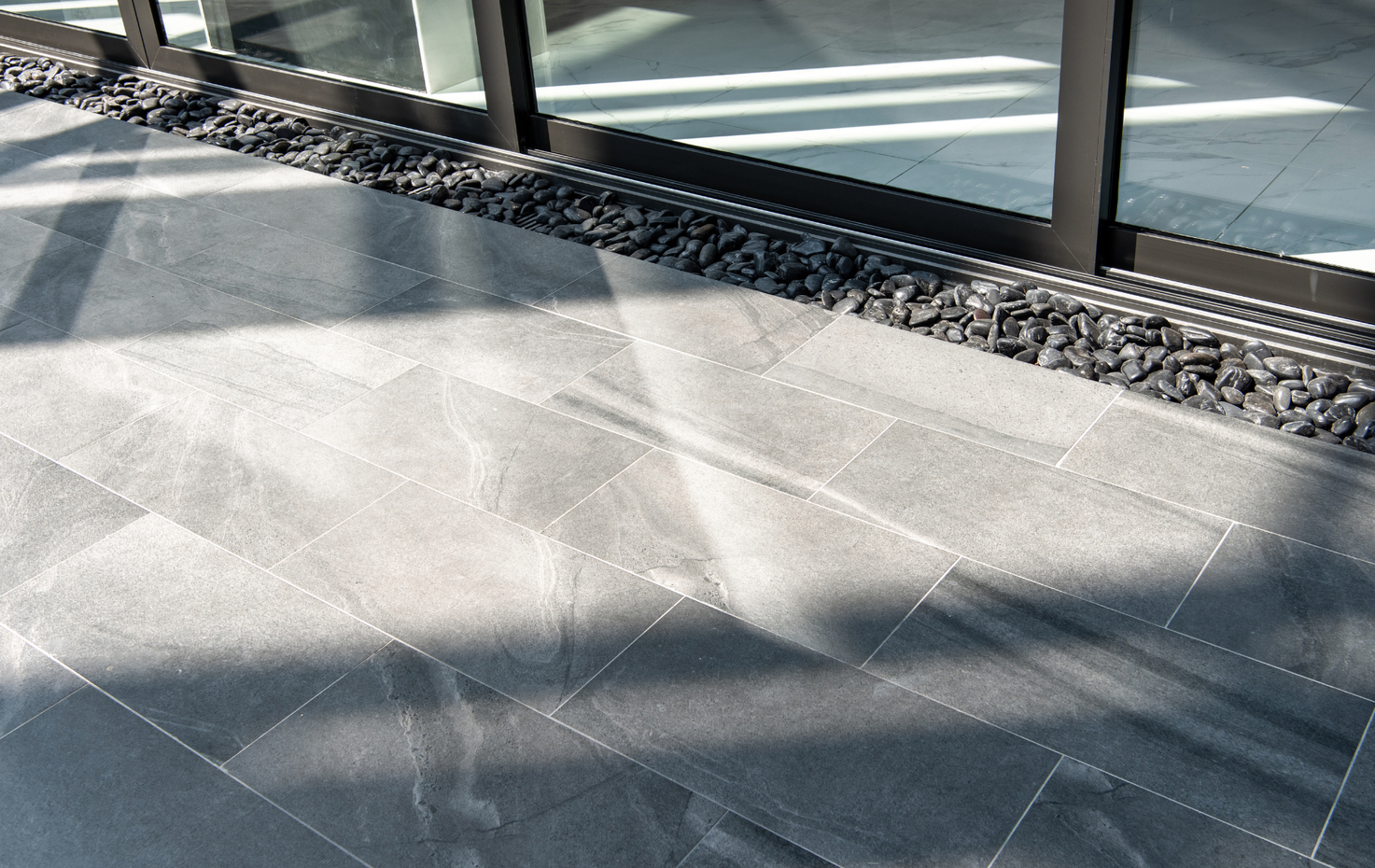 Gray tiled floor with dark pebbles along the base of a glass wall.