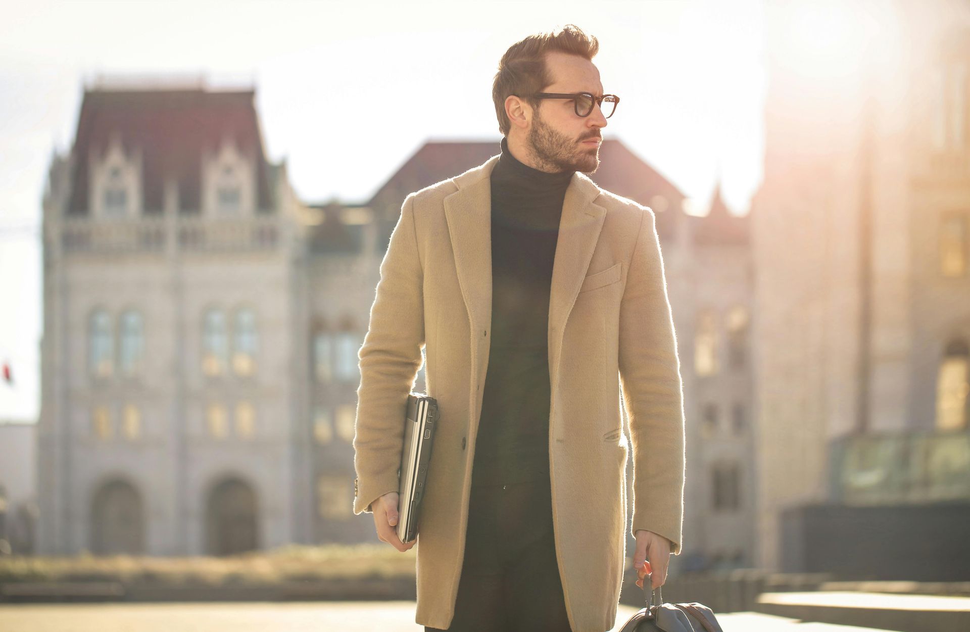 A man in a tan coat is holding a briefcase and a laptop.