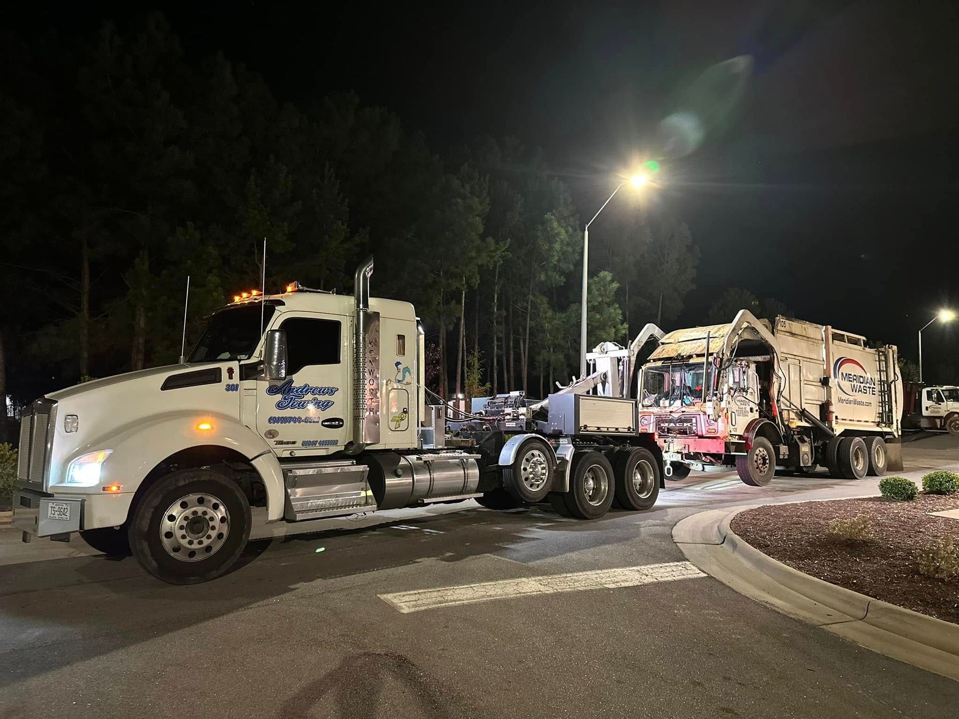 Two garbage trucks are parked on the side of the road at night.