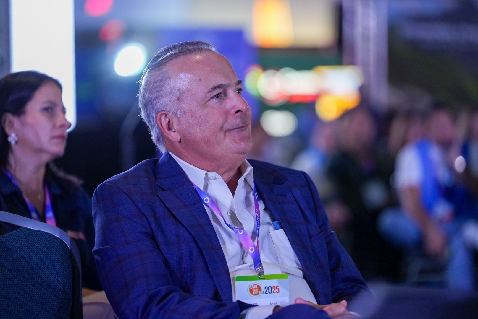 Man in a blue blazer smiles at a conference, wearing a lanyard. Woman to his left.