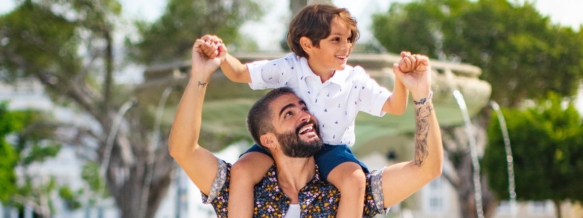 A man is carrying a little boy on his shoulders in front of a fountain.