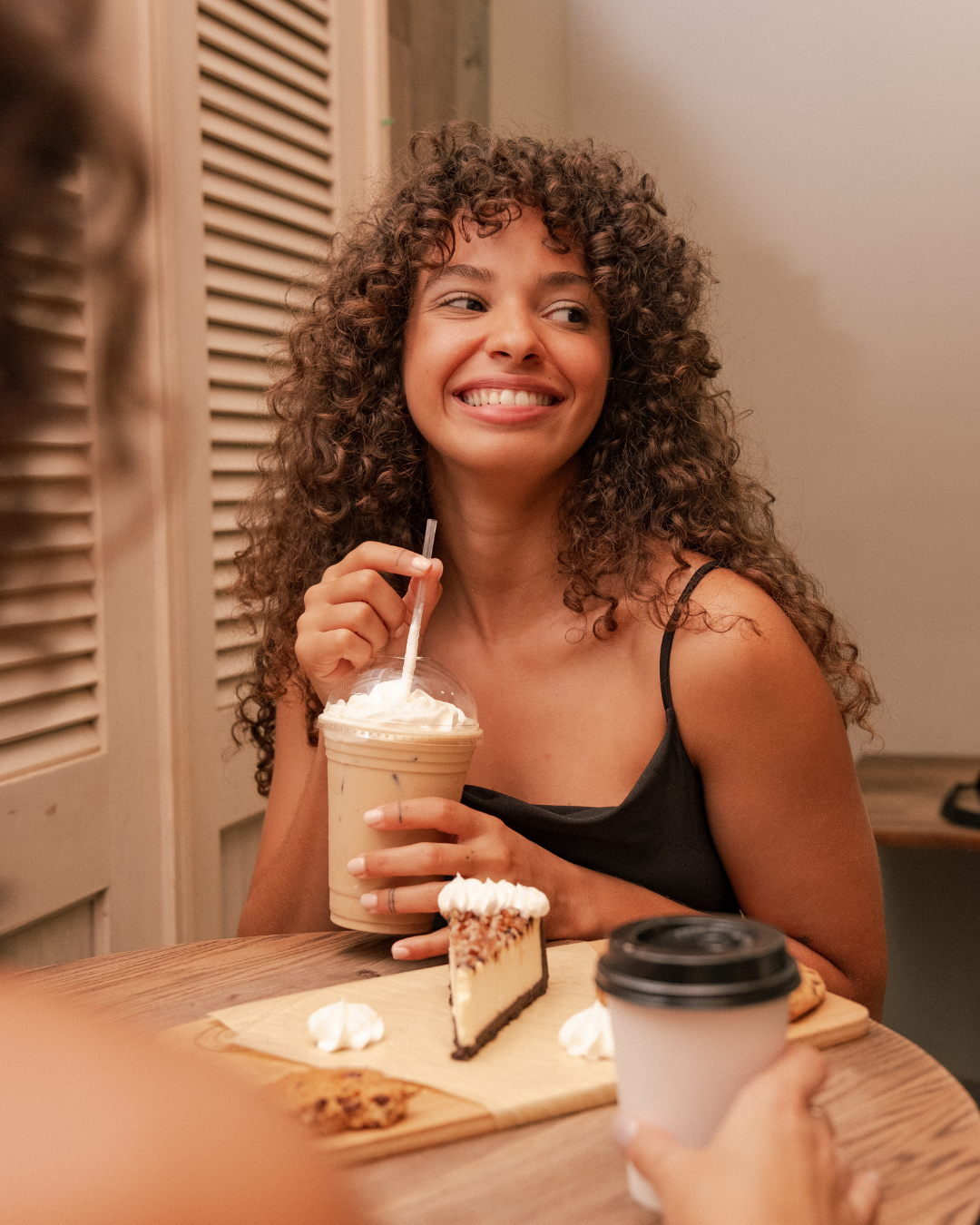 A curly-haired person smiling at a companion while holding an iced coffee at a cafe table with a slice of cheesecake.