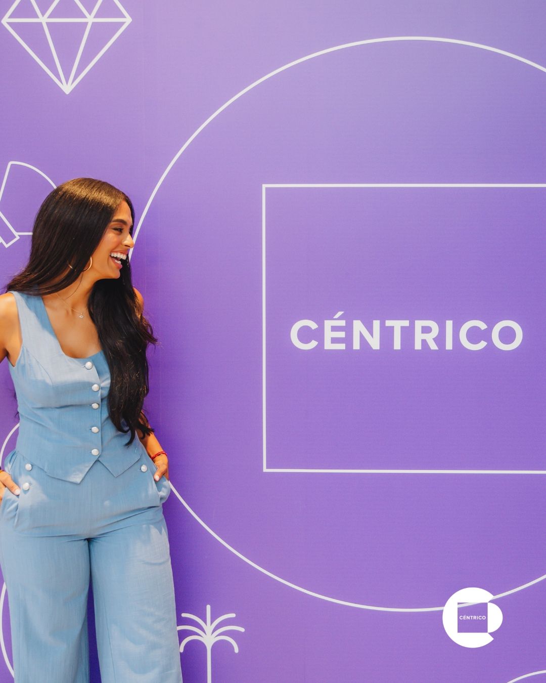 Woman in blue outfit standing by a purple CÉNTRICO backdrop with geometric graphics and a smile