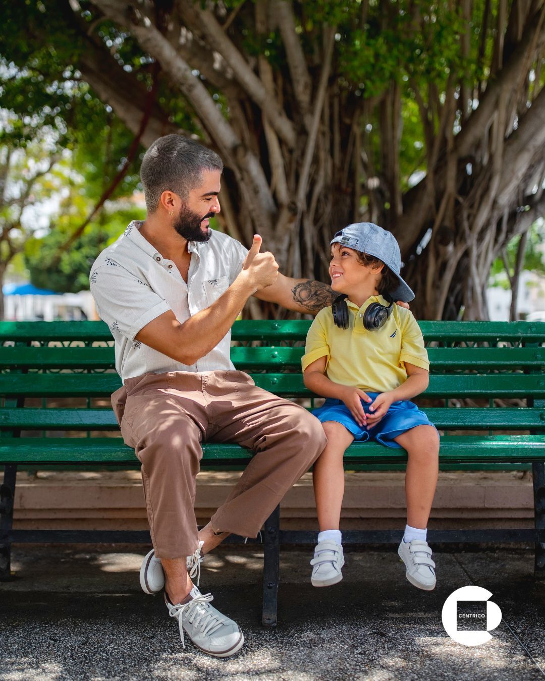 Man and child sitting on a green park bench, smiling and gesturing with hands under a tree.