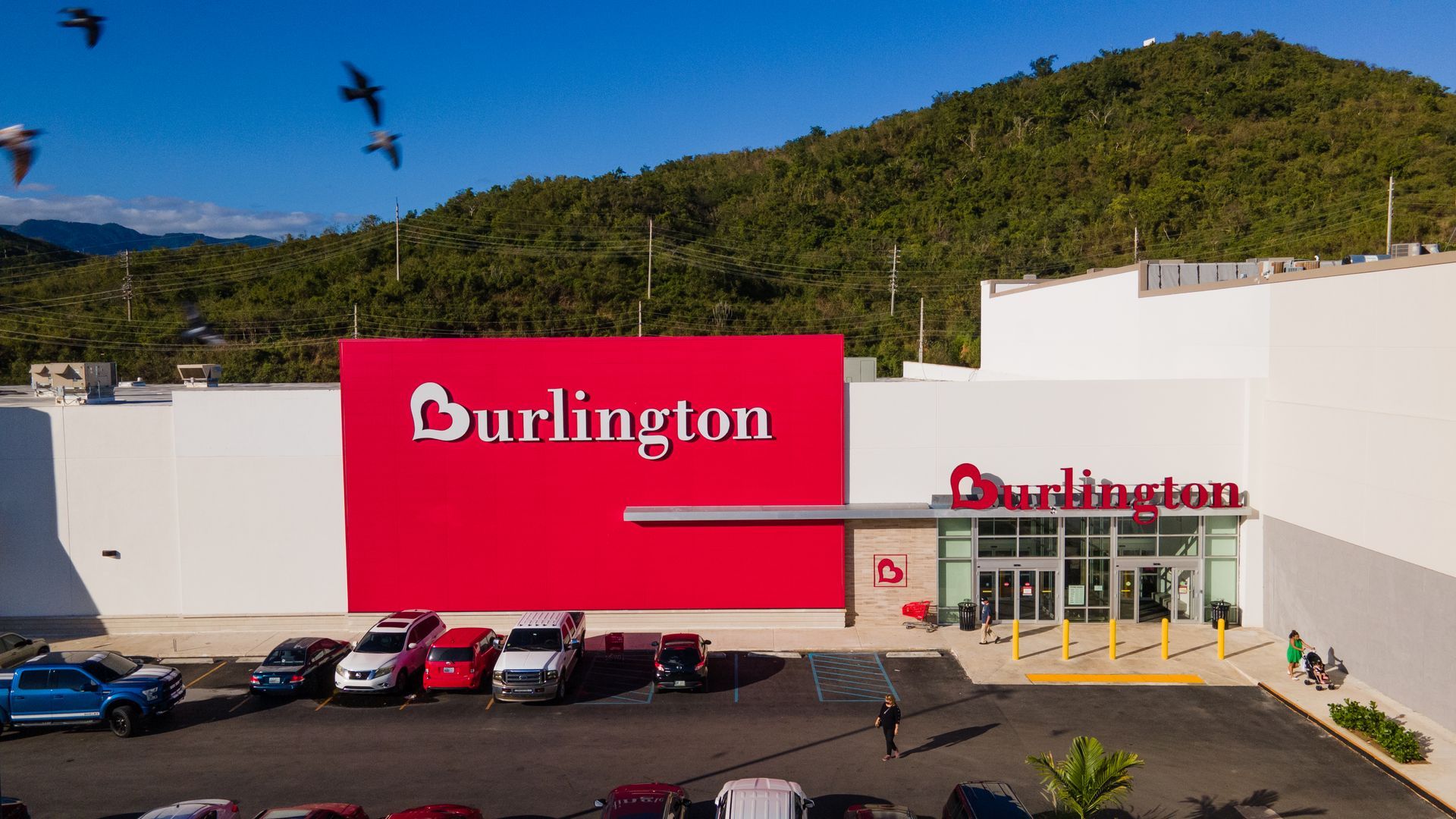Entrance to a Burlington store with a red exterior and illuminated sign. Customers can be seen shopping inside.