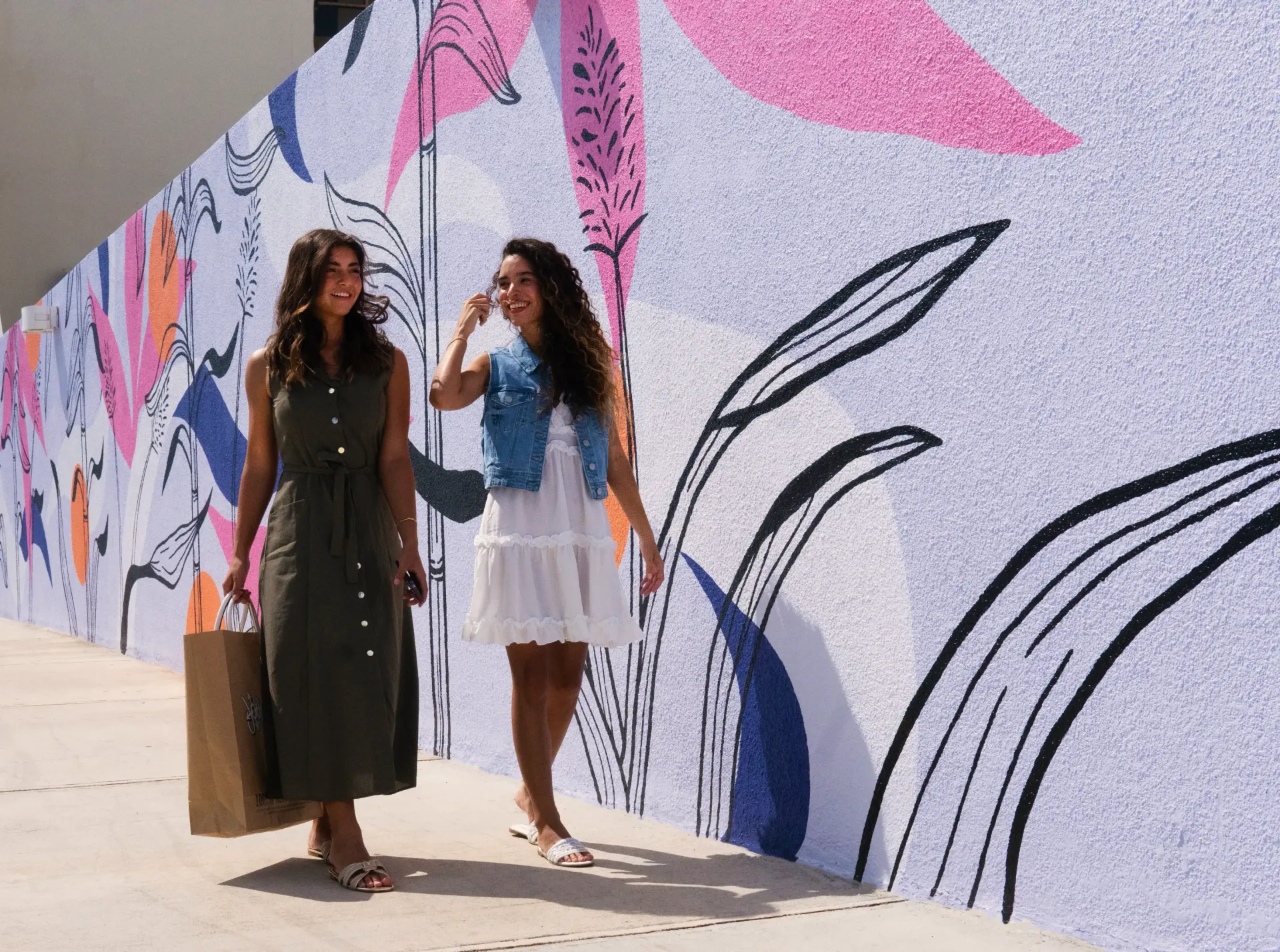 Two women are walking down a sidewalk in front of a mural.