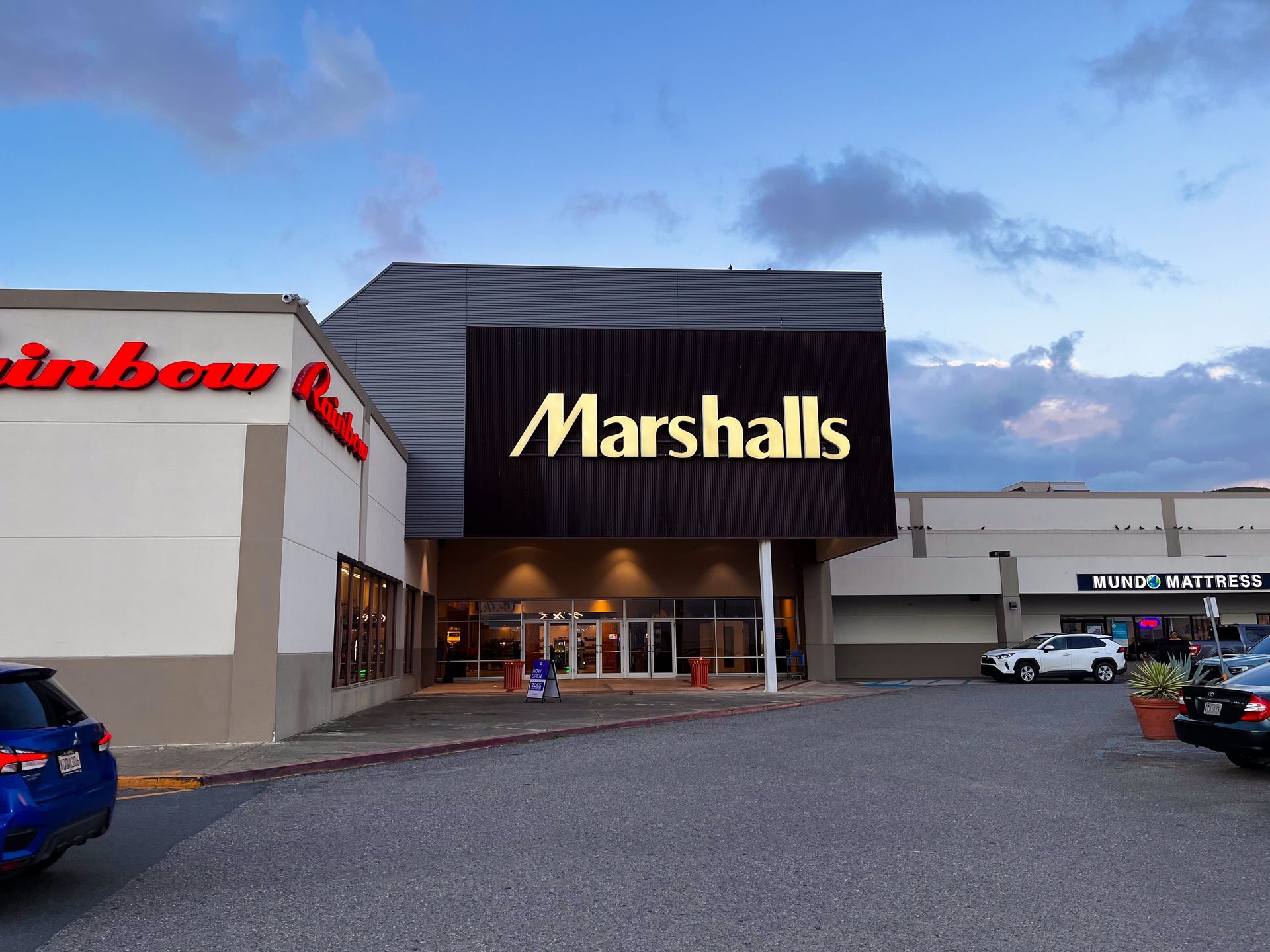 Exterior view of a Marshalls and Rainbow store on a cloudy day, with signage and a gravel parking lot.