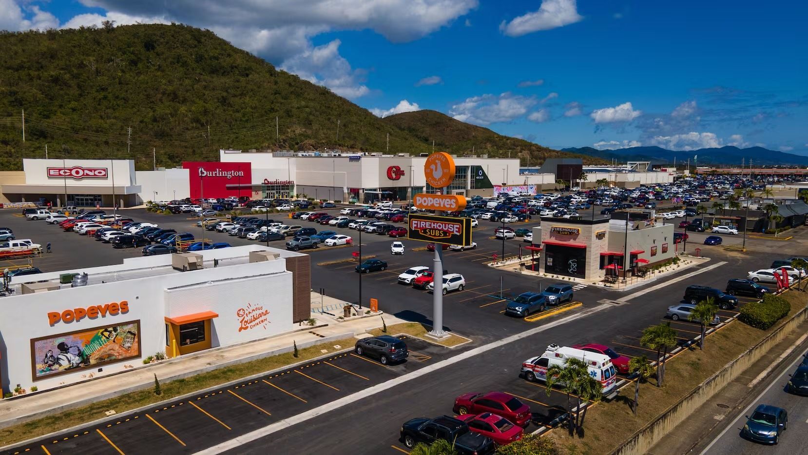 Aerial view of a shopping center with various stores, a busy parking lot, and mountains in the background.