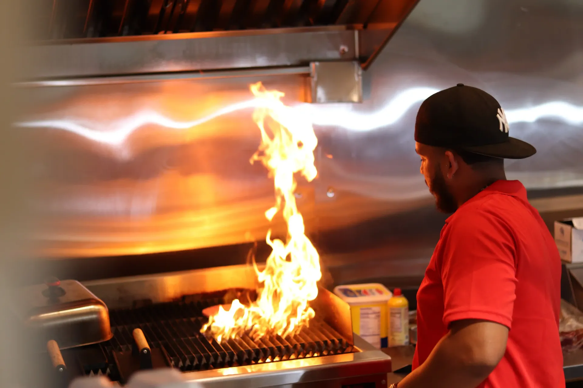 Chef in red shirt watches flames erupt from a grill in a restaurant kitchen.